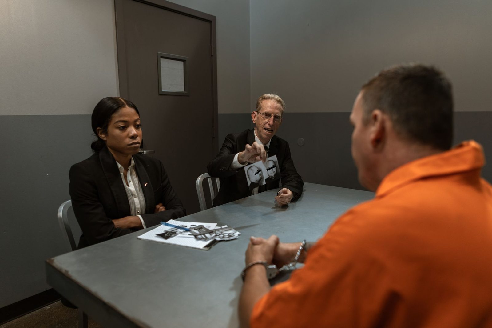 A person in an orange jumpsuit sits across from two detectives in a dimly lit interrogation room.