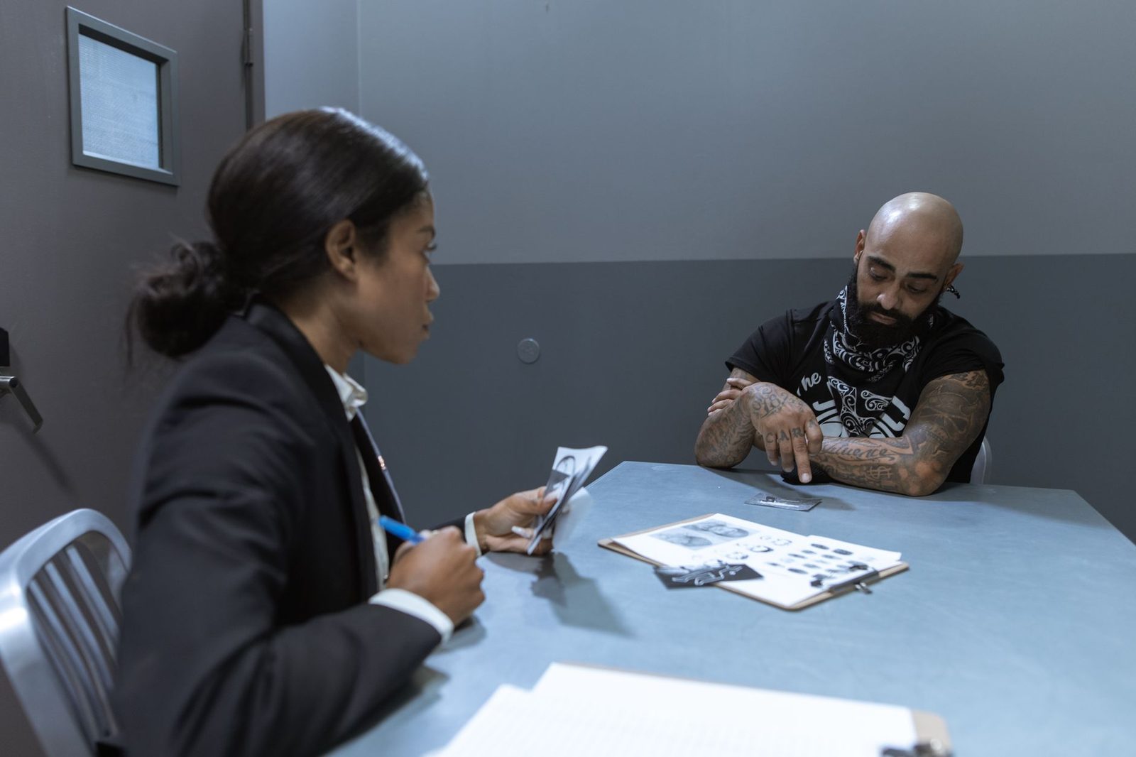 Police officer questioning a seated suspect across a table covered with case files and evidence in an interview room.
