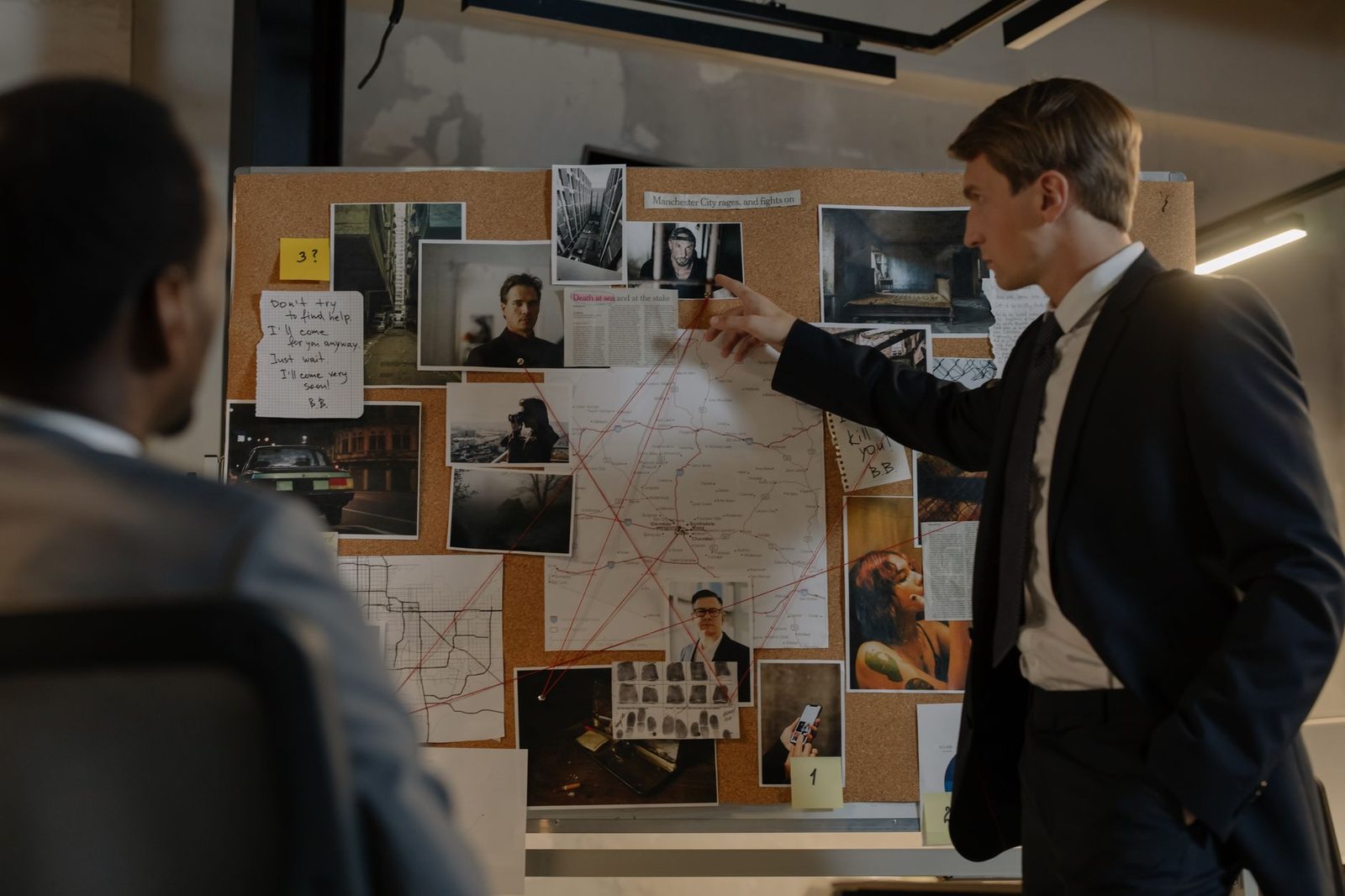 Two detectives review suspects and evidence pinned to a cork board in an office.